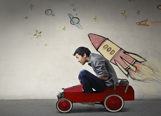The portrait of a young Asian guy in a red toy car in front of a gray wall with spaceship and planets.
